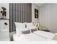 Modernes Schlafzimmer mit zwei zusammengestellten Einzelbetten, weißer Bettwäsche und einem hohen, gepolsterten Kopfteil aus grauen Stoffpaneelen. Helle Wände mit Zierleisten, Wandlampen, ein gerahmtes Bild sowie eine kleine Sitzbank mit grünen Kissen ergänzen den Raum.