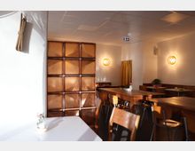 Innenansicht eines Cafés mit schlichter Einrichtung, Holzstühlen und -tischen. Die weißen Wände sind mit warmem Licht von wandmontierten Leuchten beleuchtet, und ein Raumteiler aus Glasbausteinen trennt den Bereich teilweise ab.