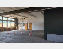 Ein leerer industrieller Innenraum mit Betonboden, großen Fensterfronten und weißen Wänden. Im Vordergrund stehen farbige Absperrkegel, und an der Wand sind zwei orangefarbene Türen sowie technische Installationen sichtbar.