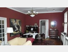 Wohnzimmer mit roten Wänden, einem Esstisch mit Stühlen in der Mitte, einem Klavier an der Wand und einer Kommode mit einem Spiegel daneben. An den Wänden hängen Kunstwerke, und ein großes Fenster lässt Tageslicht herein.