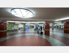 Unterirdischer Bahnhofsvorplatz mit roten und grauen Bodenfliesen, mehreren Geschäften entlang der Wände und einer kreisförmigen, hellen Deckenbeleuchtung. Zwei Personen mit Koffern gehen in Richtung eines Rollbands, das zu den Bahngleisen führt.