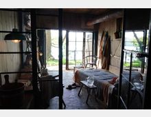 Rustikales Zimmer mit Holzmöbeln und natürlichen Materialien, darunter ein Bett mit gestreiftem Überwurf, Hocker als Nachttische und hängende Kleidungsstücke an der Wand. Tageslicht fällt durch große Glastüren ein, die einen Blick auf einen grünen Garten mit Bäumen und See im Hintergrund ermöglichen.