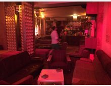 Innenraum einer Bar mit gedämpfter Beleuchtung in rot-orangen Tönen. Der Raum ist mit Vintage-Sofas und Sesseln möbliert, die um kleine Tische gruppiert sind, und im Hintergrund befindet sich eine Theke mit Flaschen und Dekorationen.