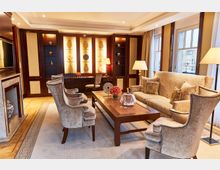 Elegantes Wohnzimmer in warmen Farbtönen mit einer großen Sofagruppe, zwei Sesseln, einem Couchtisch und einem Kamin. Im Hintergrund eine beleuchtete Holz-Wandvitrine mit dekorativen Objekten, daneben Fenster mit cremefarbenen Vorhängen und Stehlampen.