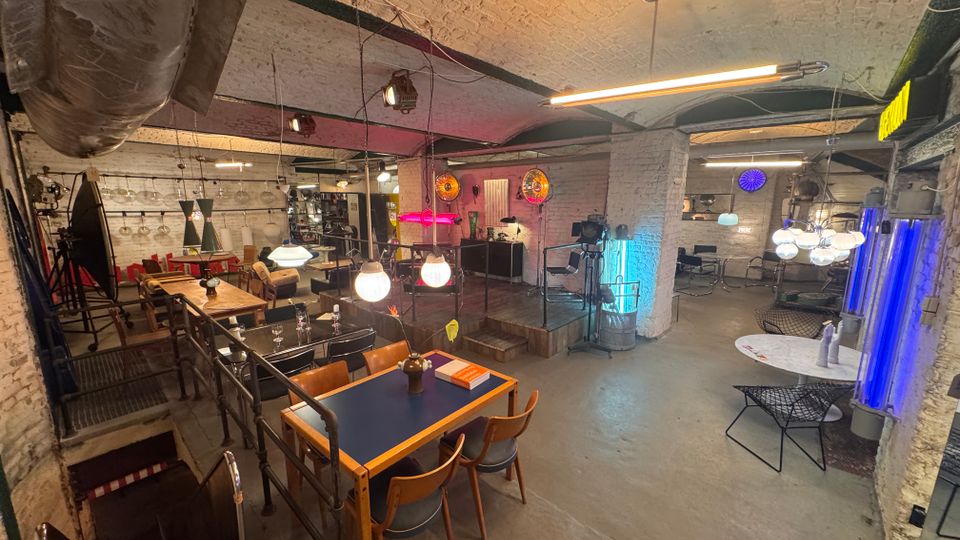 Industriell gestalteter Innenraum mit weiß gestrichenen Ziegelwänden, offener Decke und sichtbaren Metallträgern. Der Raum ist mit einer Mischung aus Holztischen, Stühlen und modernen Leuchten ausgestattet, ergänzt durch farbige Neonlichter und Metallgeländer.