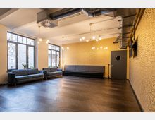 Ein moderner Lounge-Bereich mit Holzboden, goldener Wandverkleidung und mehreren Sitzgelegenheiten, darunter grau gepolsterte Sofas und eine lange Bank. Große Fenster lassen Tageslicht herein, während mehrere stilvolle Deckenleuchten und sichtbare Lüftungsrohre das industrielle Design unterstreichen.