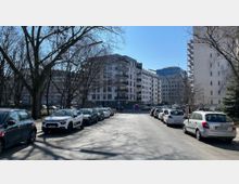 Straßenansicht in einer urbanen Umgebung mit parkenden Autos zu beiden Seiten. Im Hintergrund sind moderne Wohn- und Bürogebäude zu sehen, umliegende Bäume ohne Laub deuten auf die Winter- oder Frühjahrszeit hin.