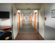 Ein langer, heller Korridor in einem Gebäude mit weißen Wänden und orangefarbenen Türrahmen. Rechts und links befinden sich mehrere Türen, am Ende des Flurs ist eine Glastür zu sehen.