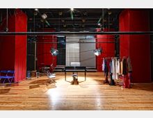 Bühnenraum mit Holzboden, roten Wänden und Beleuchtungseinrichtungen an der Decke. In der Mitte stehen ein Klavier und ein Hocker, daneben Kleiderständer mit Kostümen sowie blaue Klappstühle.