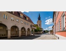 Straßenszene in einer kleinen Stadt mit historischen Gebäuden, darunter ein helles Gebäude mit Arkaden links und eine Kirche mit Turmuhr und spitzer Turmhaube im Hintergrund. Die Straße ist von Bäumen gesäumt, und der Himmel ist klar mit einigen Wolken.