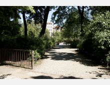 Gepflasterter Weg in einem Park, umgeben von dichten Büschen und hohen Bäumen, mit einem Metallgeländer im Vordergrund; im Hintergrund sind mehrstöckige Gebäude mit roten Dächern zu sehen.