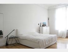 Ein schlichtes Schlafzimmer mit weiß gestrichenen Wänden, einem weißen Bett, einem kleinen Beistelltisch mit Lampe und einem weißen Schrank. Ein großes Fenster mit halbtransparenten Vorhängen lässt Tageslicht herein, und der Boden besteht aus hellen Holzdielen.