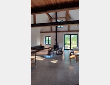 Offener Wohnraum mit hohen Decken, sichtbaren Holzbalken und großen Fenstern. Der Raum enthält eine Sitzecke mit Sofa, einen Kamin, einen Esstisch mit Stühlen und eine Glastür mit Blick ins Grüne.