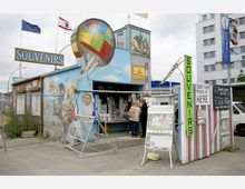 Ein kleiner Souvenirladen mit bemalter Fassade, die eine bunte Heißluftballon-Szene zeigt. Vor dem Laden stehen diverse Schilder, eine gezeichnete Stadtkarte und ein Verkaufsstand mit Postkarten; darüber wehen die Europaflagge und eine Stadtflagge.