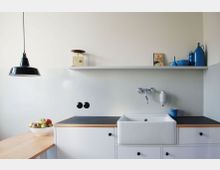 Minimalistische Küche mit weißen Wänden und schwarzen Akzenten. Zu sehen sind eine weiße Spüle mit verchromter Wandarmatur, eine Arbeitsplatte mit Holzdetails, ein Regal mit blauen Keramikdekorationen und eine schwarze Hängelampe; auf einem Tisch steht eine Schale mit Äpfeln.