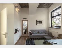 Moderner, minimalistisch eingerichteter Raum mit einem grauen Sofa, dekorativen Kissen und einem kleinen Beistelltisch. Ein großes Fenster lässt Tageslicht herein, und eine Zimmerpflanze steht auf der Fensterbank; an der Wand hängen ein gerahmtes Schwarz-Weiß-Foto und ein hölzern gerahmter Spiegel.