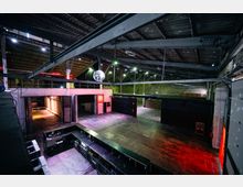 Großer, industrieller Innenraum einer Halle mit hohen Decken aus Metallträgern, rohem Betonboden und teilweise rohen Backsteinwänden. Der Raum ist spärlich beleuchtet mit roten und grünen Lichtakzenten, eine große Discokugel hängt von der Decke, und im Vordergrund ist ein langer Tresenbereich zu sehen.