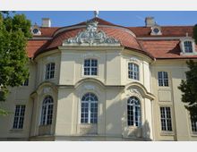 Fassade eines historischen Gebäudes mit hellgelbem Putz, gebogenen Architekturformen und weißen Verzierungen über den Fenstern. Das Dach ist mit roten Ziegeln gedeckt, und Bäume rahmen den Rand der Ansicht ein.