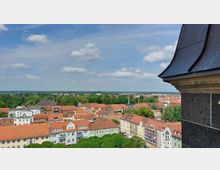 Ausblick auf eine Altstadt mit roten Ziegeldächern und mehrstöckigen, bunt gestrichenen Gebäuden, umgeben von viel Grün und Wäldern in der Ferne. Ein Teil eines Kirchturms oder Gebäudes ist rechts im Vordergrund sichtbar, der Himmel ist leicht bewölkt.