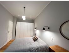 Schlafzimmer mit grauen Wänden, einem Doppelbett mit grauer Bettdecke und zwei Wandlampen darüber. Im Hintergrund steht ein großer, weißer Kleiderschrank, und rechts ist ein runder Spiegel an der Wand angebracht. Der Boden ist aus Holz.