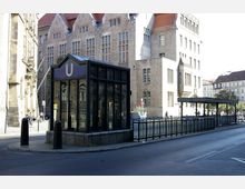 Straßenansicht mit einem Eingang zur U-Bahn, gekennzeichnet durch ein Glashäuschen mit einem großen "U" darüber. Im Hintergrund ist ein historisches Gebäude mit Sandsteinfassade und roten Dachziegeln zu sehen, flankiert von weiteren Gebäuden und einer Straße mit Fußgängern und Fahrrädern.