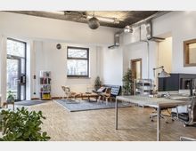 Modernes Büro mit großem offenen Grundriss, Holzboden und hohen Decken mit sichtbaren Lüftungsrohren. Der Raum ist in einen Arbeitsbereich mit Schreibtisch und Bildschirm und eine Sitzecke mit Sofa, Stühlen und Teppich unterteilt; große Fenster lassen viel Tageslicht herein.