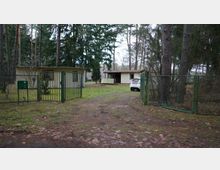 Zwei kleine, helle Gebäude in einem bewaldeten Grundstück, umgeben von einem Metallzaun mit offenem Tor. Ein weißes Auto steht vor einem der Gebäude, und der Boden ist mit Gras und Herbstlaub bedeckt.