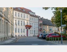 Straßenszene mit historischen Gebäuden in einer kleinen Stadt: Mehrstöckige Fassaden mit hellen Putzfarben und roten Ziegeldächern säumen eine gepflasterte Straße, vor der Autos geparkt sind. Rechts im Bild befinden sich Bäume und ein Straßenschild mit der Aufschrift „Paul-Feller-Straße“.