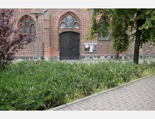 Backsteinfassade einer Kirche mit gotischen Spitzbogenfenstern und einer großen Holztür mit kunstvollen Schmiedeeisen-Verzierungen. Im Vordergrund befinden sich grüne Sträucher, ein gepflasterter Gehweg und ein Baum auf der rechten Seite.