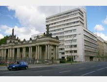 Historisches Eingangsportal mit Säulen und Skulpturen im Vordergrund, dahinter ein neoklassizistisches Verwaltungsgebäude mit mehreren Etagen und rechteckigen Fenstern. Die Szenerie liegt an einer Straße mit geparktem blauen Auto.