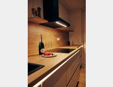 Moderne Küche mit heller Holzarbeitsplatte, integriertem Induktionskochfeld und minimalistischen Schränken. Auf der Arbeitsplatte stehen eine Sektflasche, zwei Gläser und ein Teller mit Erdbeeren und Pralinen, während eine schwarze Dunstabzugshaube mit Beleuchtung darüber montiert ist.