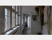 Helle Innenaufnahme eines gefliesten Flures mit weißen Wänden und großen Fenstern auf der linken Seite. Rechts hängen Pflanzen von der Decke, während eine Tür am Ende des Flures in einen weiteren Raum führt.