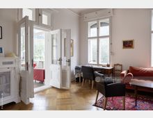 Heller Wohnraum in einem Altbau mit hohen Decken, großen weißen Flügeltüren und Parkettboden. Der Raum ist mit klassischen Holzmöbeln, einem roten Sofa und Teppich, sowie großen Fenstern mit Blick ins Grüne ausgestattet.