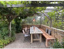 Überdachter Sitzbereich im Freien mit Holztisch und gepolsterten Stühlen sowie einer gemauerten Sitzbank. Die Pergola ist mit Weinreben und Kletterpflanzen bewachsen, umgeben von einem Garten mit viel Grün.