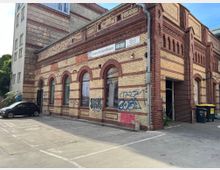 Backsteinbau mit historischer Fassade, verziert mit roten und gelben Klinkern sowie Rundbogenfenstern. Vor dem Gebäude befinden sich ein Parkplatz, ein Müllcontainer und vereinzelt Graffitis an den Wänden.