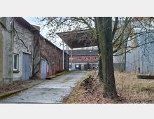 Schmaler betonierter Zufahrtsweg in einem ehemaligen Industriehof, flankiert von alten Backstein- und Putzgebäuden mit großen Metalltüren und überwachsenen Fassaden. Rechts stehen kahle Bäume und hohes Gras neben einer Wellblechhalle; im Hintergrund sind weitere Industriegebäude und ein Vordach zu sehen.