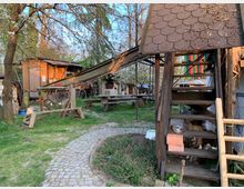 Ein rustikaler Außenbereich mit Holzbauten, darunter eine überdachte Treppe und eine kleine Hütte. Im Hintergrund sind ein Backofen, Tische und Bänke sowie blühende Bäume zu sehen, gepflasterte Wege führen durch die grüne Wiese.