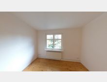 Ein leerer, heller Raum mit weiß gestrichenen Wänden, einem Holzboden und einem Fenster mit Blick ins Grüne. Unter dem Fenster befindet sich ein Heizkörper.