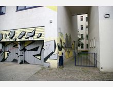 Eine schmale Gasse zwischen zwei Gebäuden mit schlichter Putzfassade; links befindet sich eine Garagenwand mit großflächigem Graffiti, rechts ein Zaun mit offenem blauen Tor. Im Hintergrund sind weitere Gebäudefassaden und ein Baum zu sehen.