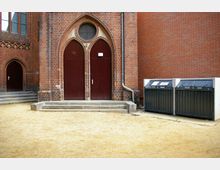 Backsteinfassade eines Gebäudes im gotischen Stil mit zwei großen, spitzbogigen Holztüren und einem runden Fenster darüber. Daneben stehen zwei Müllcontainer auf einem sandigen Boden; Regenrinnen verlaufen an der Wand.