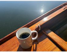 Holzboot auf ruhigem Wasser, mit glänzendem Bootsrand und Sonnenreflexionen auf der Wasseroberfläche. Im Vordergrund steht eine weiße Kaffeetasse auf einer hölzernen Sitzbank im Boot.