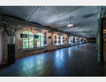 Großer, langgestreckter Innenraum mit einem industriellen Charakter, gekennzeichnet durch unverputzte Wände, ein gewölbtes Backsteindach und große, bogenförmige Fenster, durch die Tageslicht einfällt. An den Wänden sind Kunstwerke und Fotografien ausgestellt, die an Metallgestellen befestigt sind.