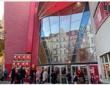 Außenansicht eines modernen Theater- oder Kinoeingangs mit einer großen Glasfassade, die umliegende Altbauten reflektiert. Links und rechts der Eingangstüren sind rote Wände mit beleuchteten Filmplakaten und Werbung angebracht; davor stehen mehrere Menschen.
