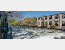 Industriegebäude aus Backstein und Beton mit zahlreichen Fenstern, entlang eines breiten Flusses mit starker Strömung; im Vordergrund ein altes Wasserrad, umgeben von Bäumen und Vegetation.