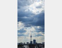 Blick auf die Berliner Skyline mit dem Fernsehturm im Zentrum, flankiert von modernen Hochhäusern und Kirchtürmen, unter einem Himmel mit dicht bewölkten und sonnigen Bereichen. Im Vordergrund sind Dächer von Wohngebäuden zu sehen.