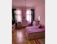 Helles Schlafzimmer mit Holzboden und hohen Decken, dekoriert mit Stuckverzierungen. Im Raum stehen ein Bett mit rosa Bettwäsche, weiße Kommoden, ein Sessel, ein kleiner Spiegel und farbige Vorhänge vor großen Fenstern, die natürliches Licht einlassen.