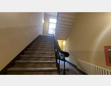 Treppenhaus in einem Wohn- oder Bürogebäude mit einer steinernen Treppe, einem schwarzen schmiedeeisernen Geländer und beige gestrichenen Wänden. Im Hintergrund ist ein großes Fenster mit Rundbogen zu sehen, das Tageslicht hereinlässt.