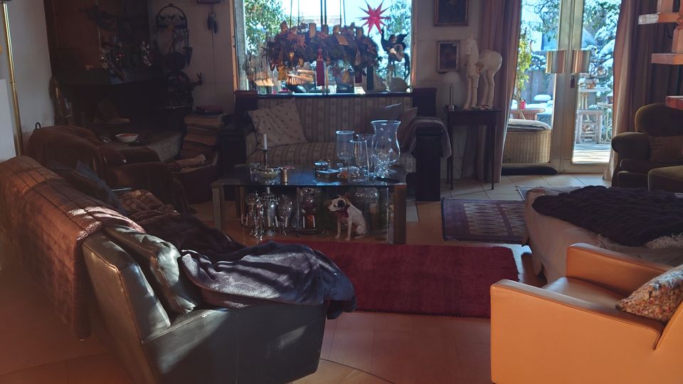 Wohnzimmer mit Holzboden und Teppich, möbliert mit Sofas und Sesseln. Im Hintergrund große Fensterfront mit Blick auf eine dekorierte Terrasse und hölzerne Treppe, die zu einer Galerie im oberen Bereich führt.