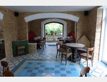 Ein gemütlicher Innenraum eines Cafés mit Wänden aus beigem Backstein, farbigen Fliesenböden und rustikalen Holzmöbeln. Im Hintergrund führt eine große, weiße Rundbogenöffnung zu einer Terrasse mit Pflanzen.