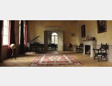 Ein historischer Salon mit antiker Einrichtung, darunter ein Flügel, ein Globus, ein Kamin und mehrere Stühle. Die Wände sind gelblich und teils abgenutzt, große Fenster mit roten Vorhängen lassen Tageslicht herein, und ein großer Teppich mit rotem Muster liegt im Mittelpunkt des Holzfußbodens.